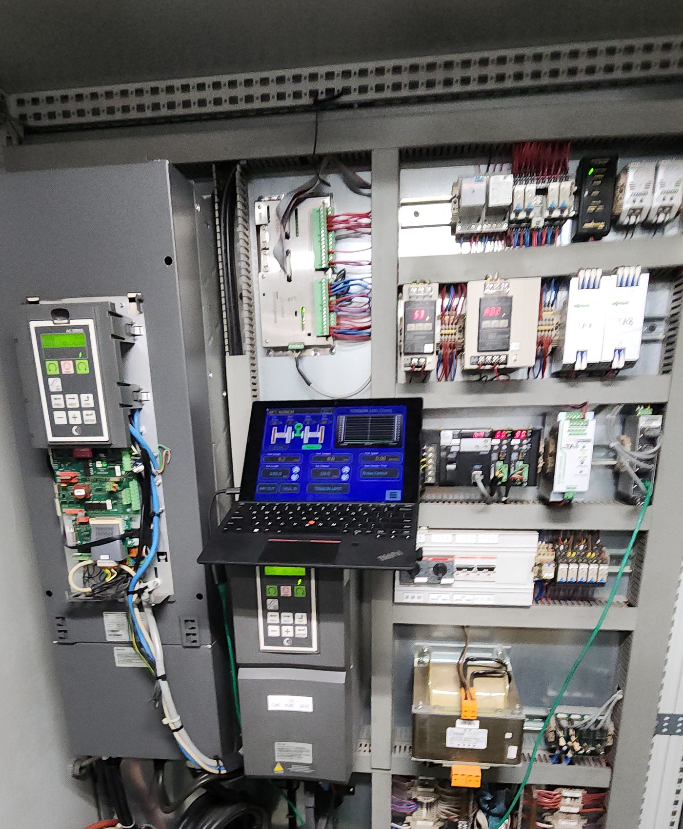 Laptop connected to open control cabinet during commissioning — PLC, motor drives, terminal blocks